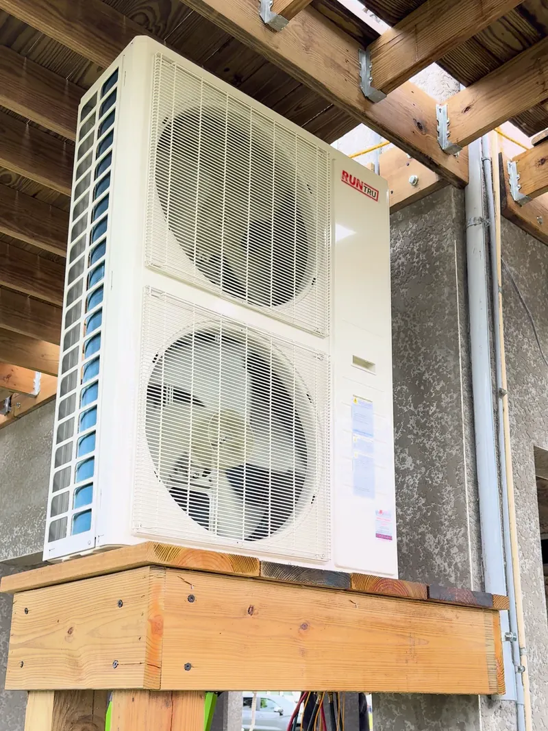 Outdoor condensing unit with two fans set on an elevated wood stand under a raised home