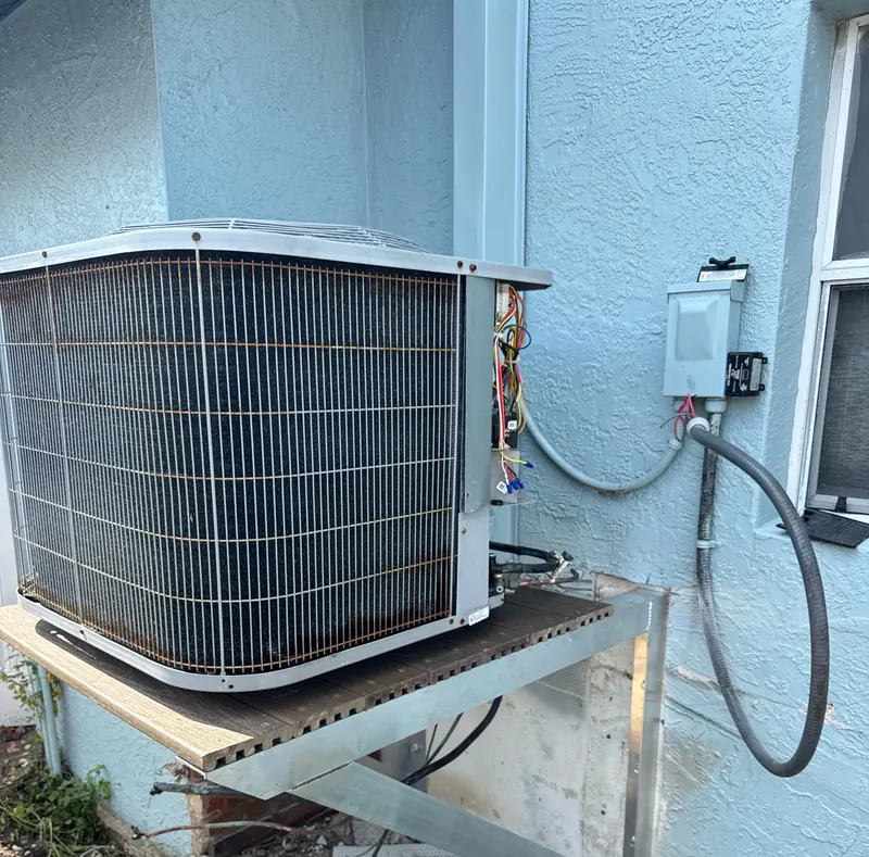 Outdoor condenser set on a wall-mounted aluminum platform beside a stucco home, with its electrical disconnect